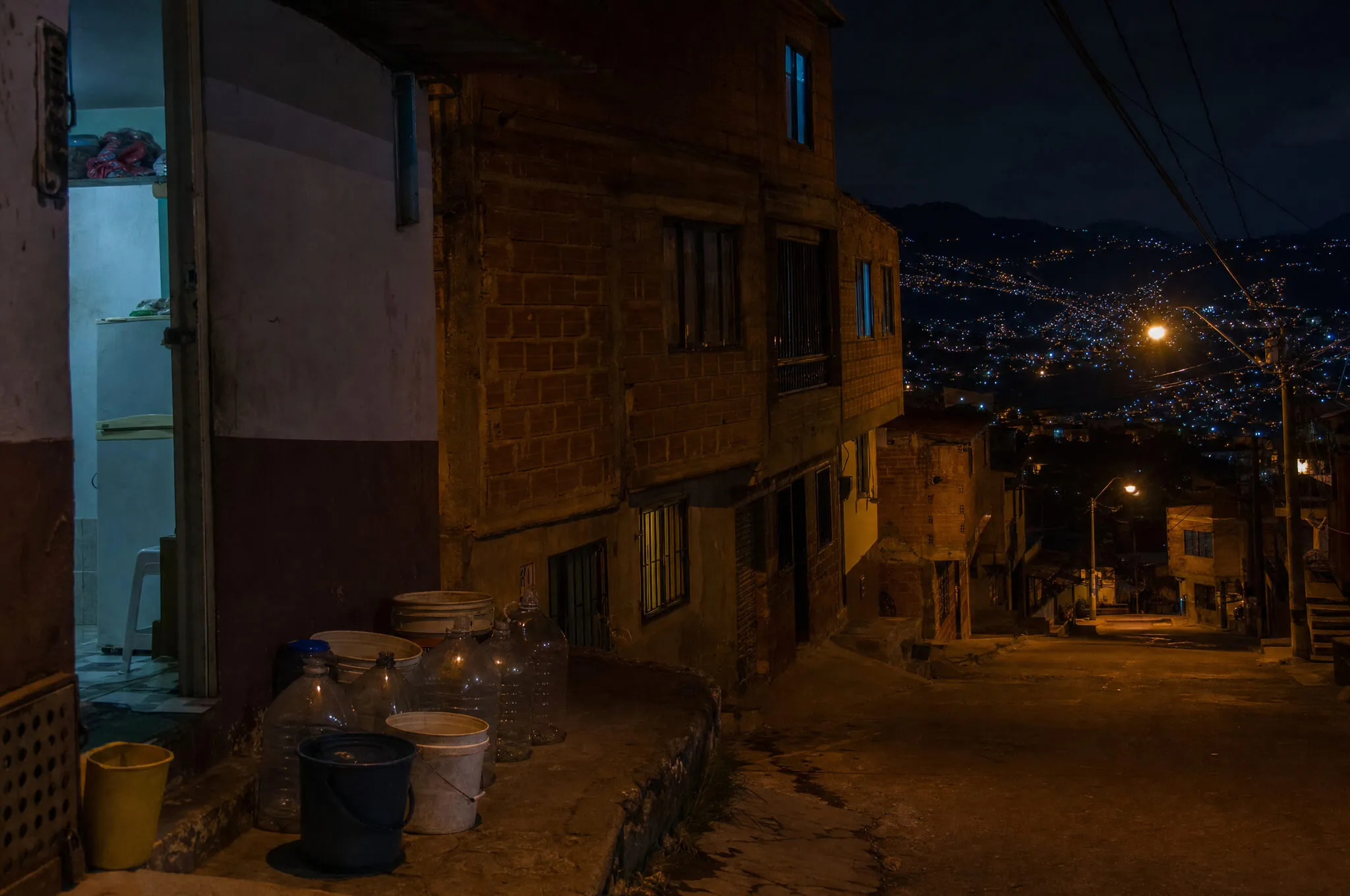 Empty water jugs and buckets lined up outside a brick apartment building on a Medellín hillside street at night.