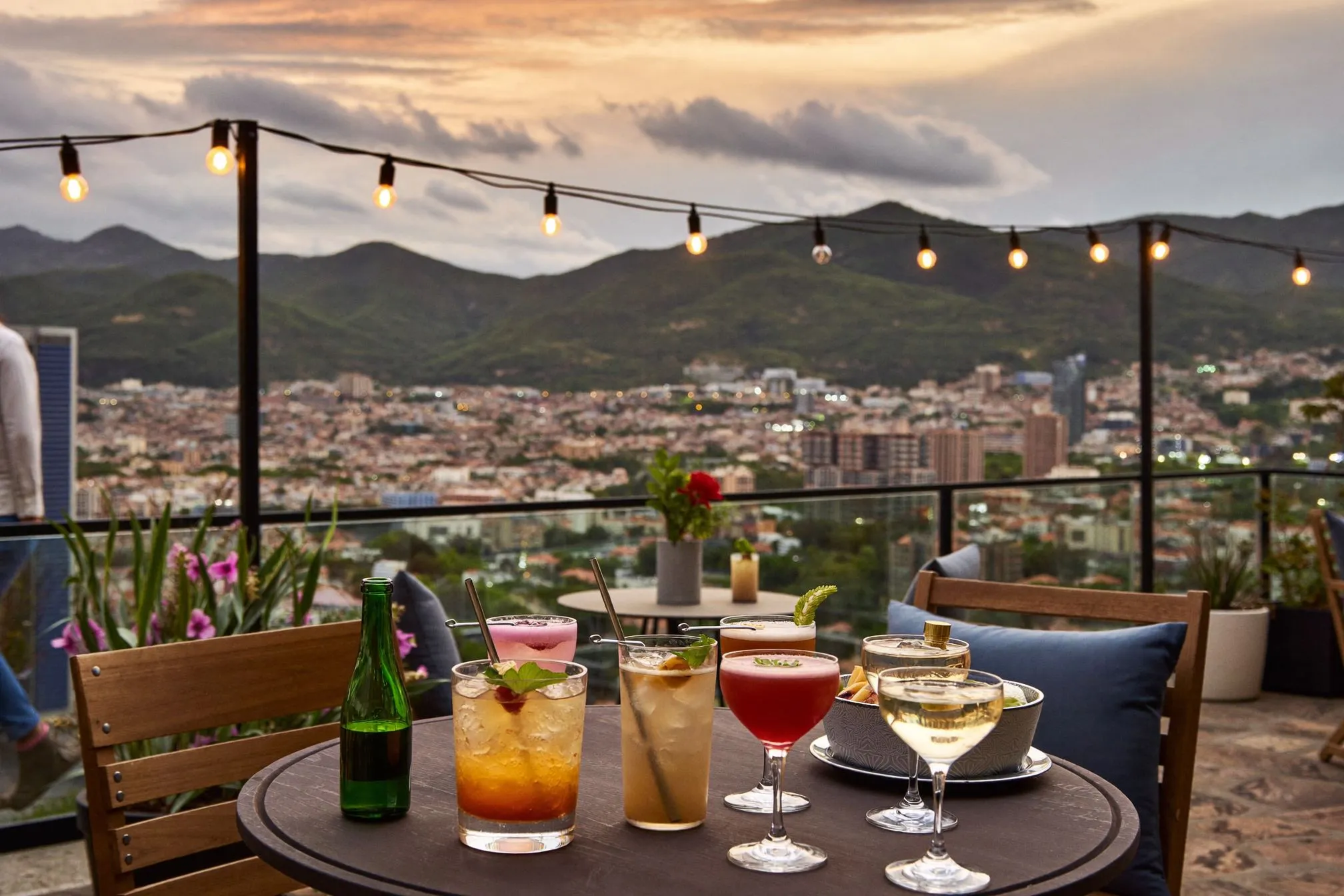 Craft cocktails on a wooden table at a rooftop bar terrace overlooking the Aburrá Valley at golden hour